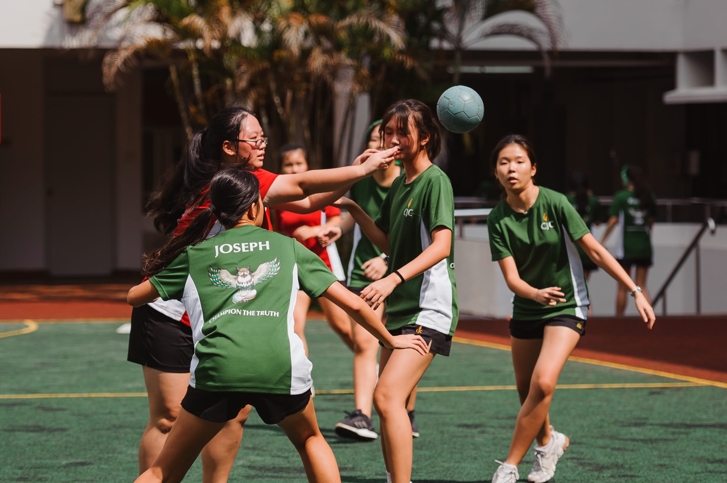 Girls in green and red jerseys play handball on an outdoor court, with a teal ball in the air.